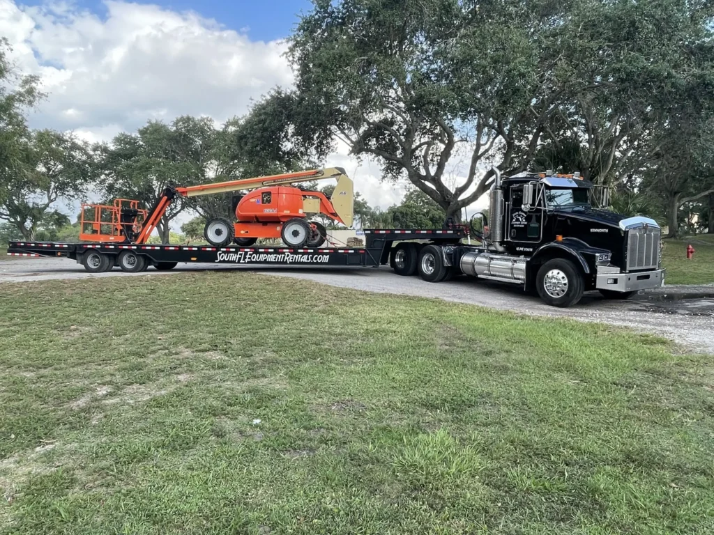 Semi-truck hauling an Equipment rental West Palm Beach flatbed trailer with an orange telescopic boom lift parked on a grassy area.