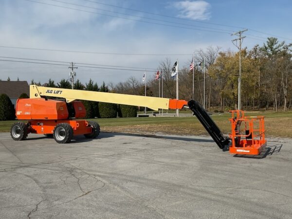 An 86ft Boom Lift with an extended arm, painted orange and yellow, is parked on an empty paved lot near grass, trees, and several flagpoles.